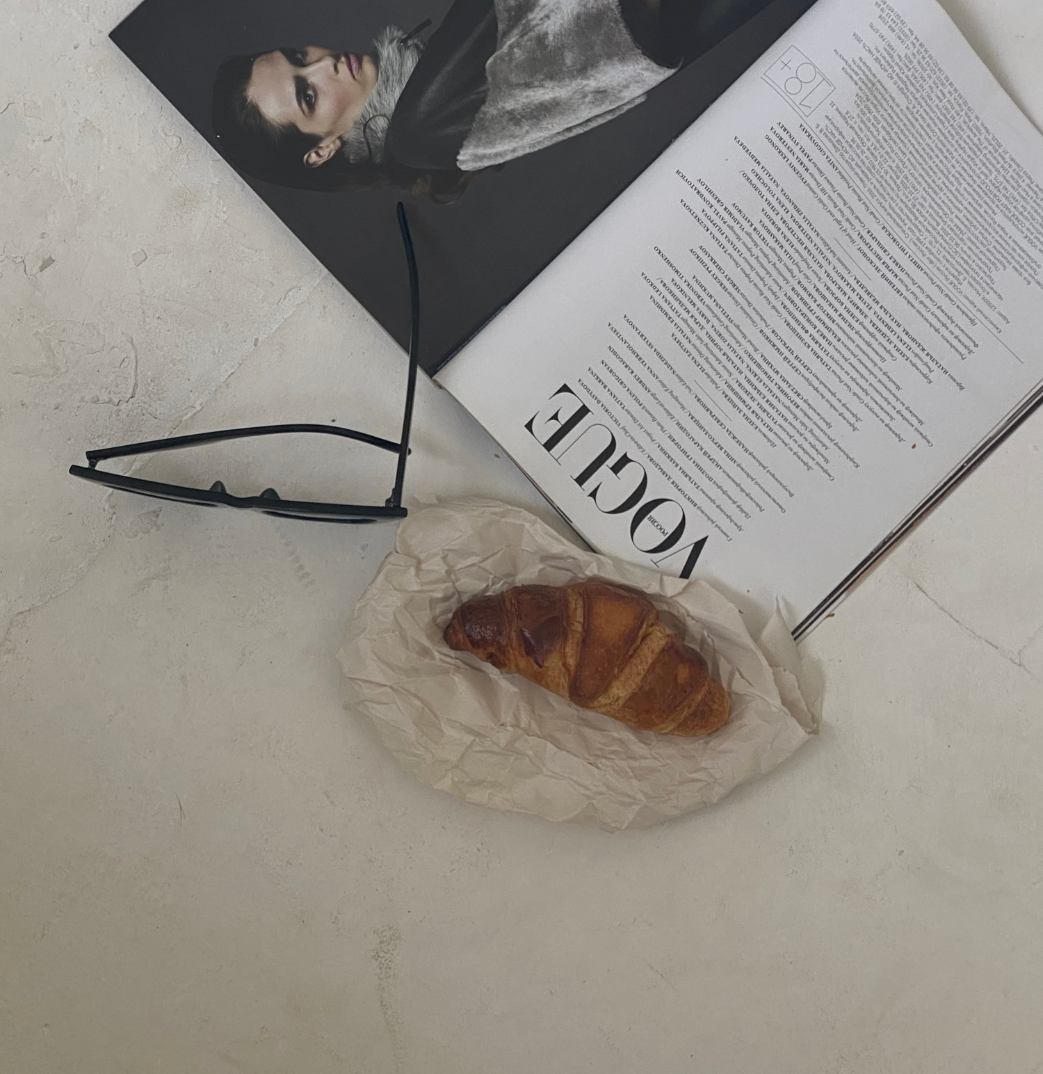 Croissant, sun glasses, and Vogue magazine on a cream textured surface