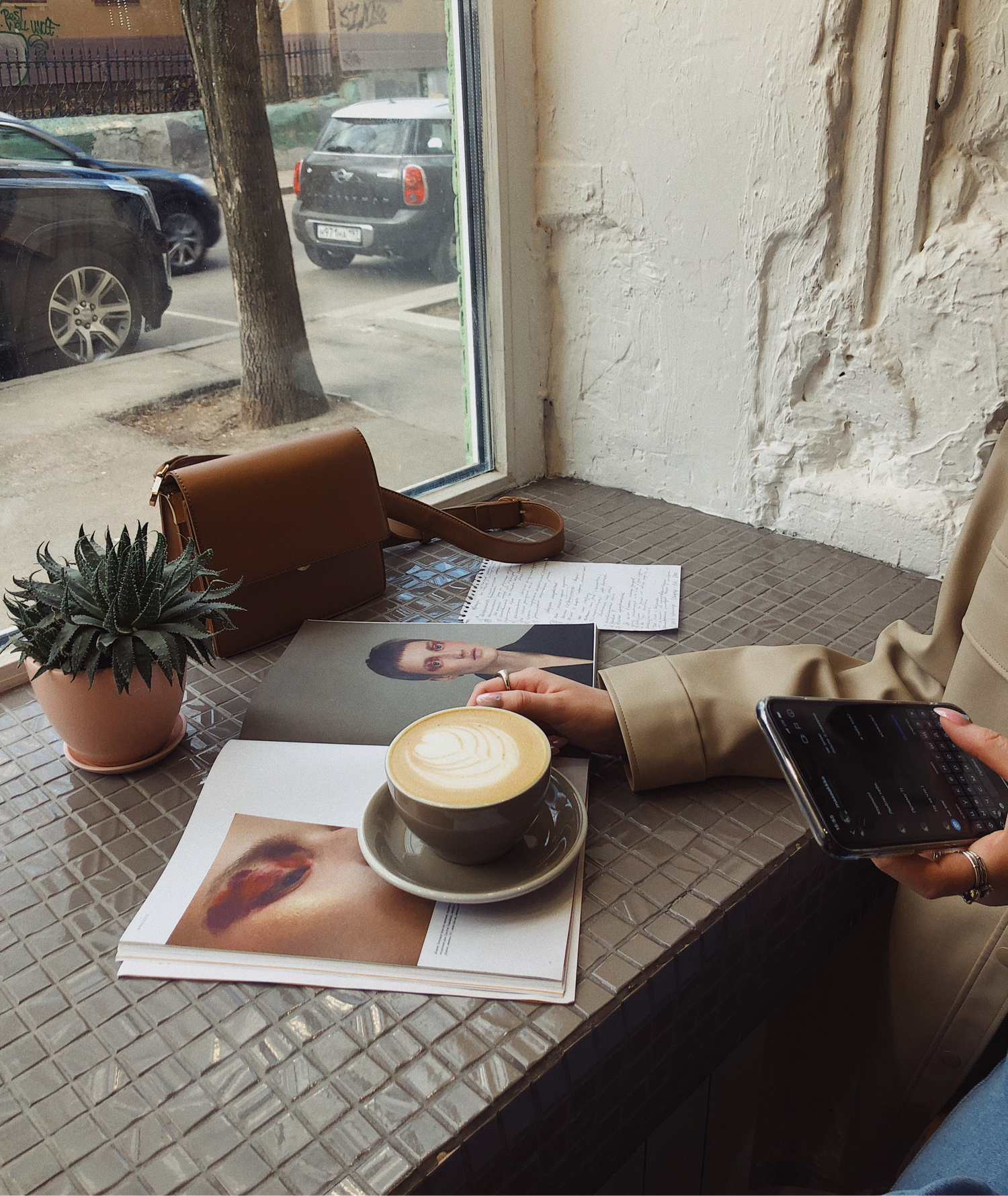 Model sitting in a cafe while on iPhone with latte, magazine, note paper, plant and purse on tiled table