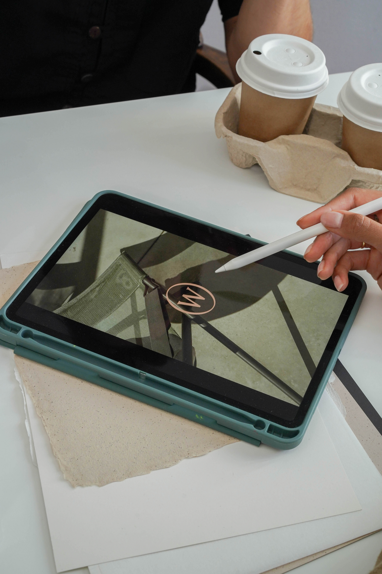 Model using an Apple Pencil on an iPad that's on a white table with pieces of paper and two coffee cups in a coffee carrier on it