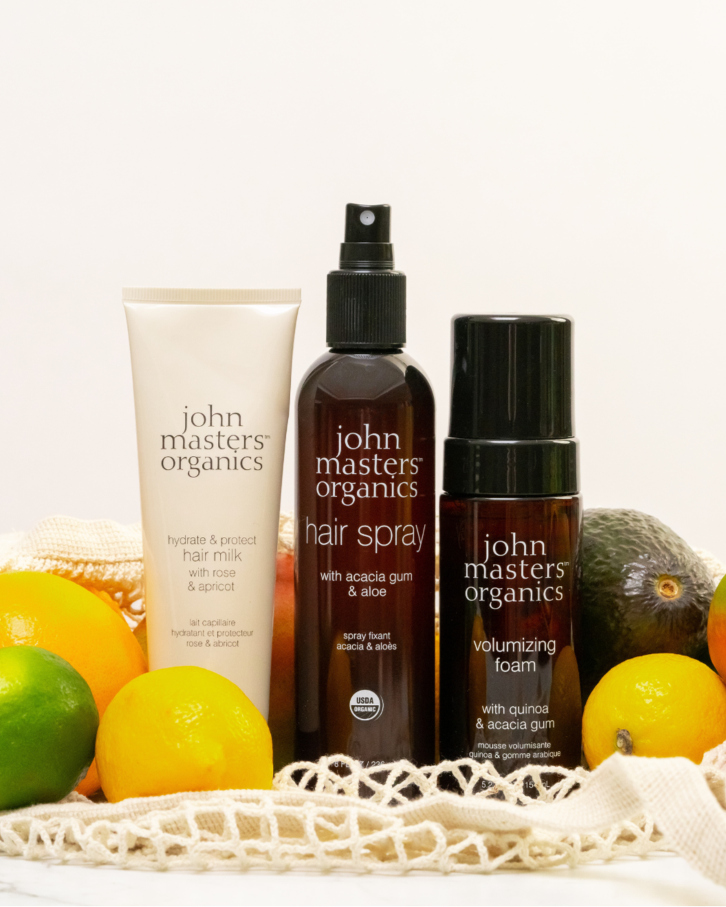 John Masters Organics Hydrate & Protect Hair Milk, Hair Spray, and Volumizing Foam with lemons, avocados, and limes on a net bag