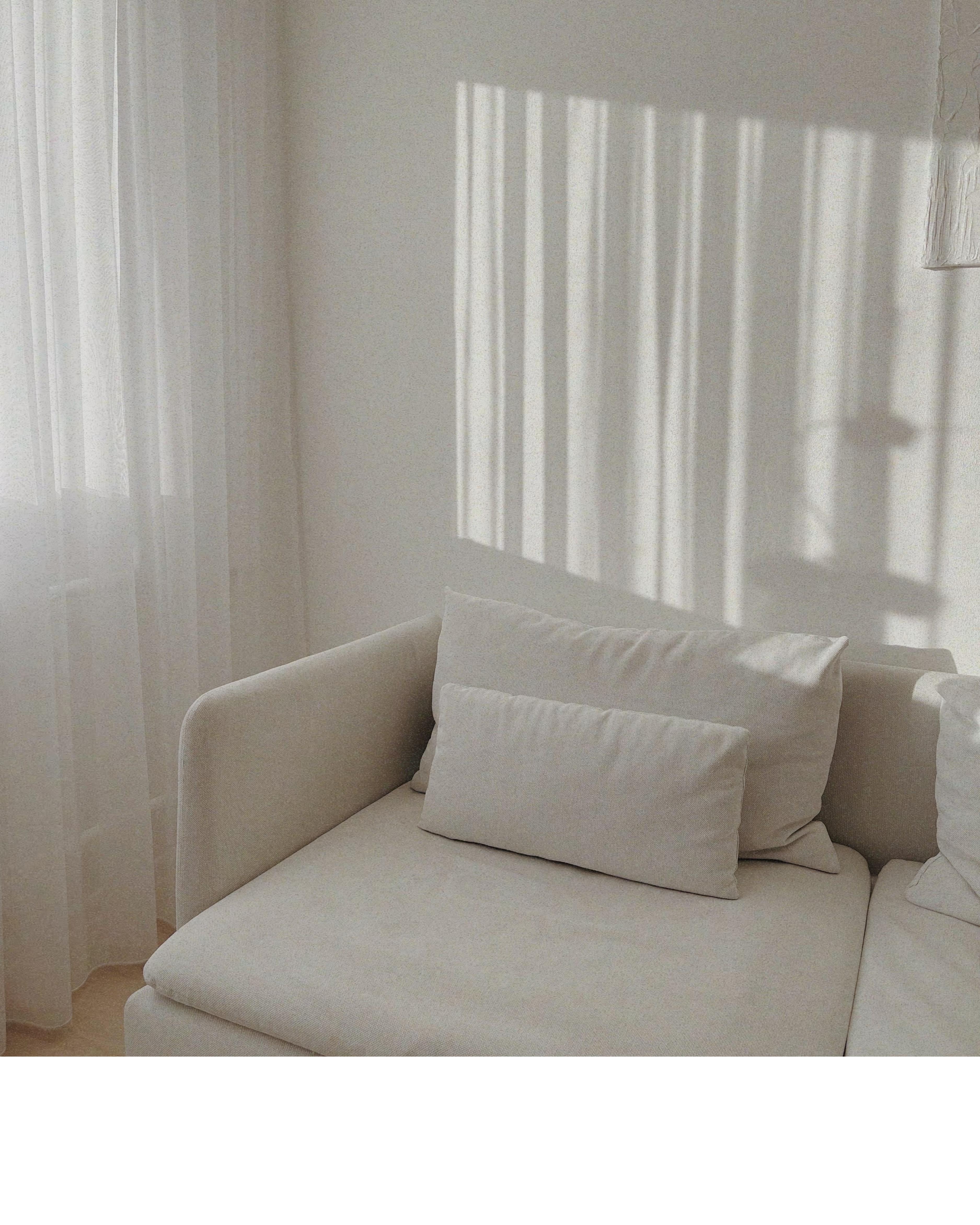 White couch in a neutral room with white curtains and sun shining through window casting aesthetic shadows on the wall