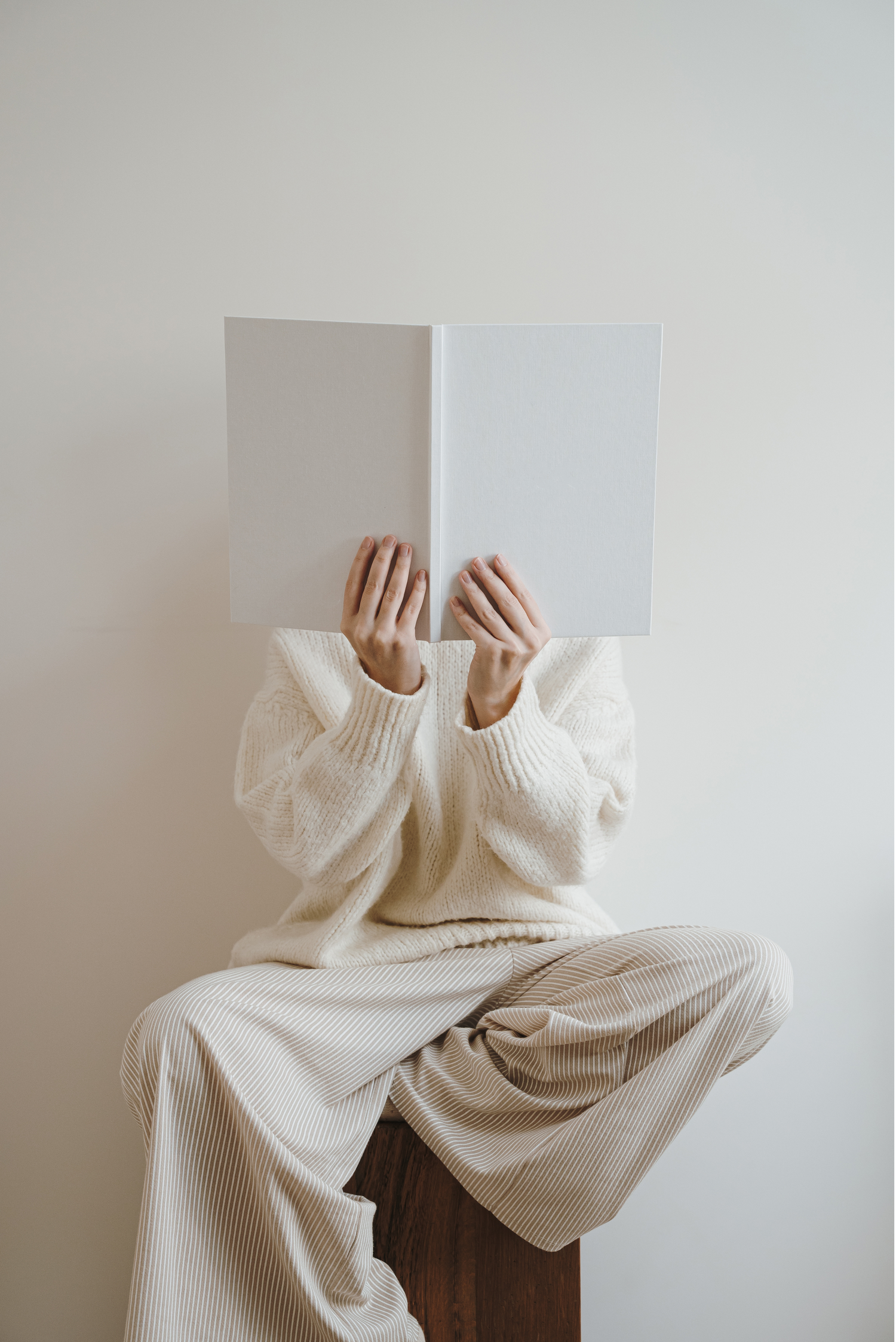 Model in all white holding a white book up in front of face while sitting on a wooden block